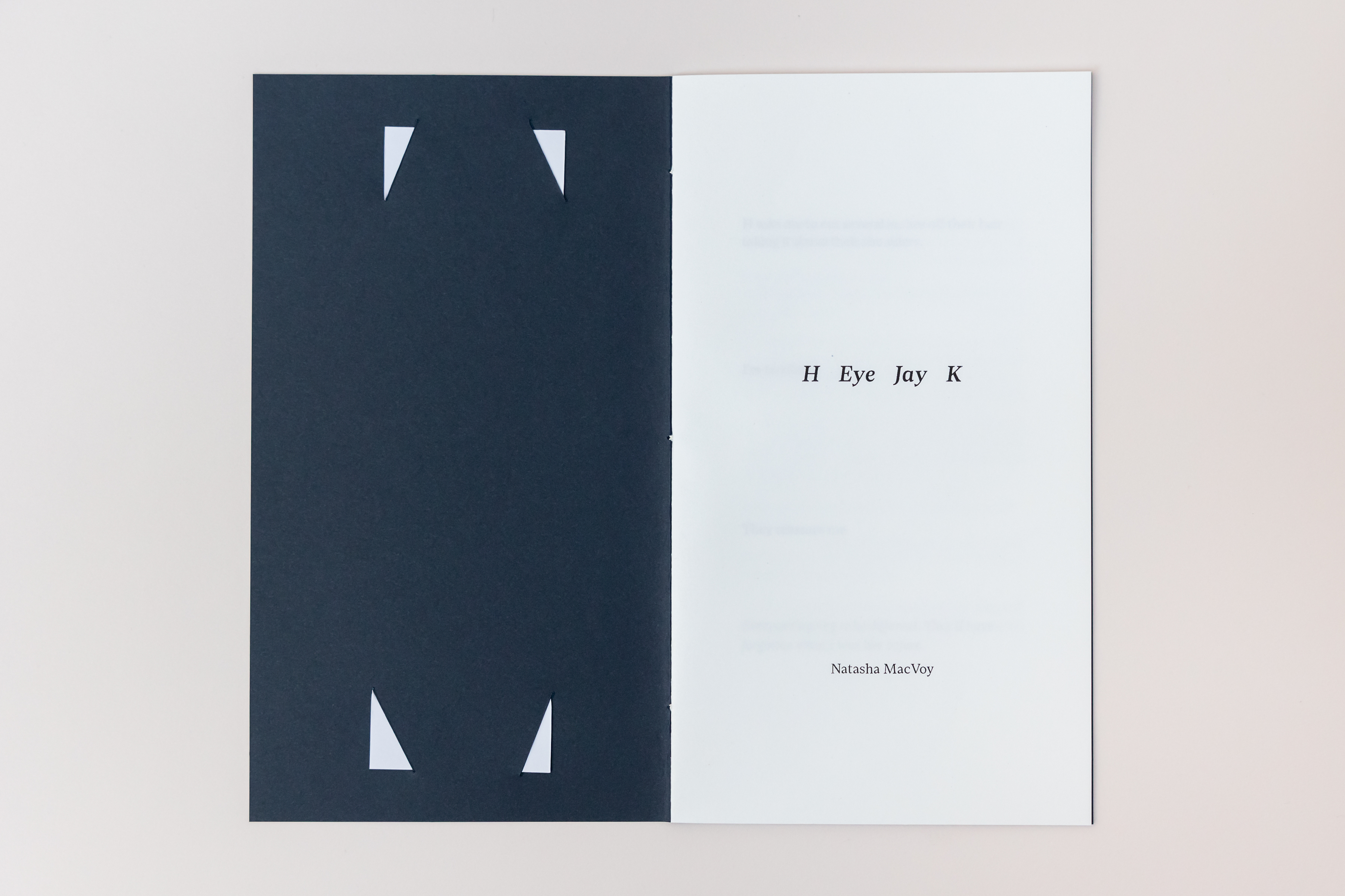 Image shows a small handmade publication open on the first internal page. It has a dark blue cover through which four small white triangles indicate the inserted bookmark from its cover. On the first page is written H Eye Jay K in the centre with Natasha MacVoy below it.