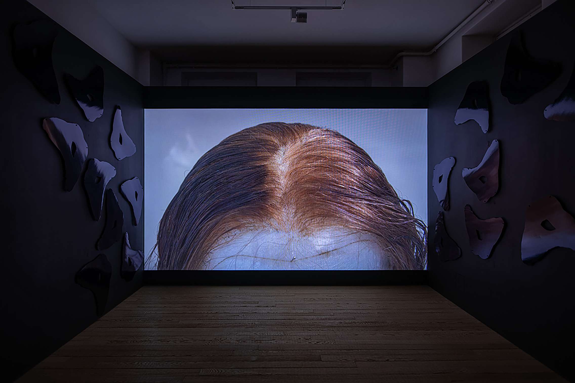 Image shows a film being projected onto, and filling, the back wall of a gallery. The film shows a wig on a head block with a pin holding it in place in the centre of the forehead. Either side of the film are dark walls with a repeating reflective shape.