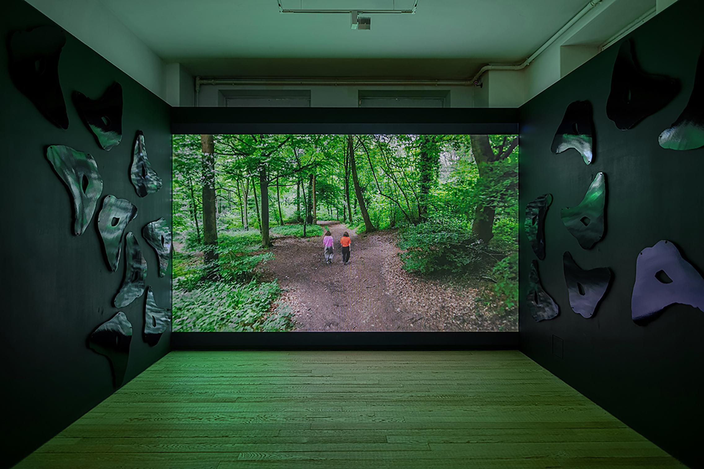 Image shows a film being projected onto, and filling, the back wall of a gallery. The film shows two figures walking away from the camera in a wood. Either side of the film are dark walls with a repeating reflective shape.