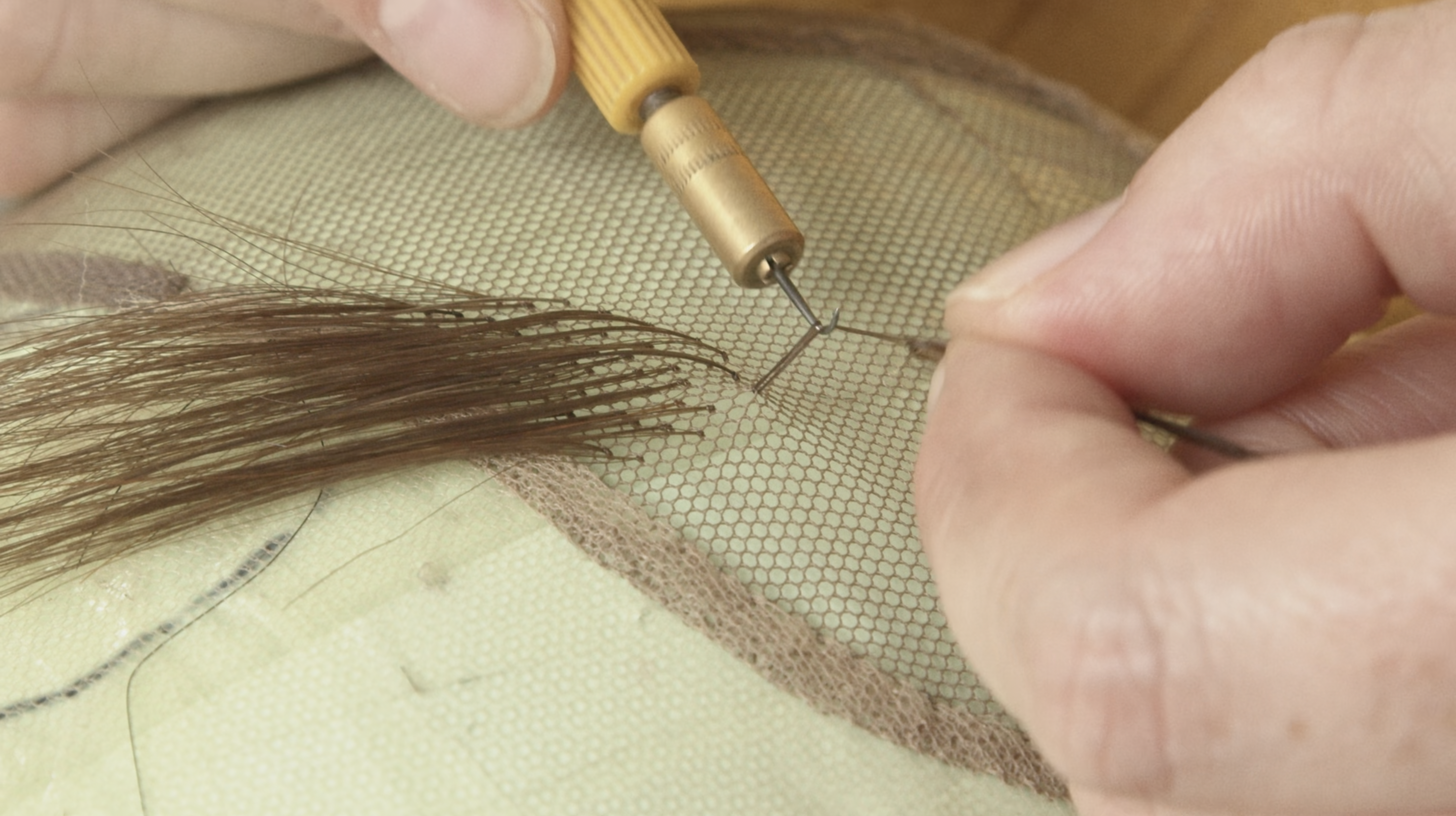 Image shows a pair of hands knotting several strands of hair through wig lace, in close detail.