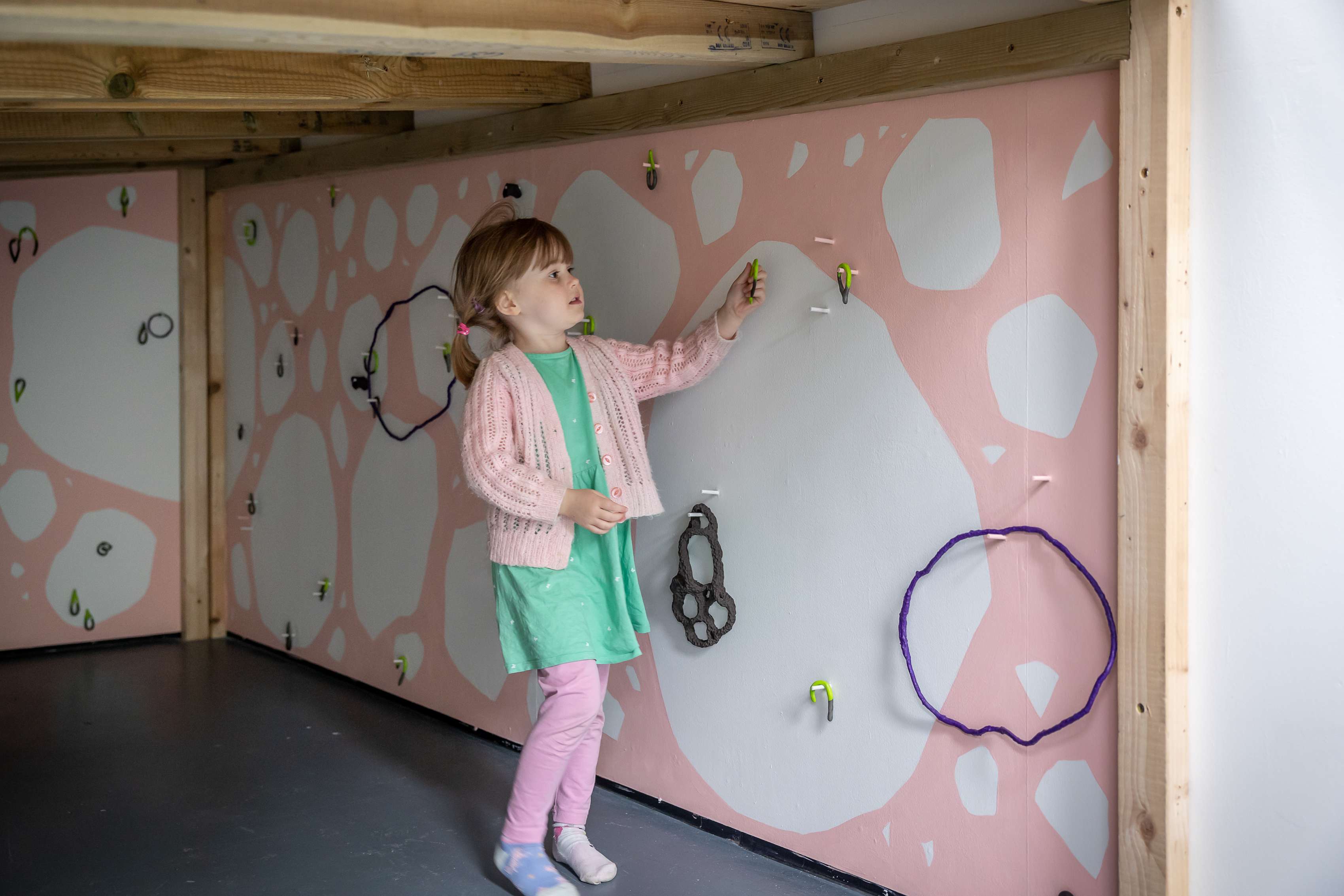 A young child holds a motif that she's lifted from a peg on the wall in front of her, which is painted pink with white boulders and rocks.