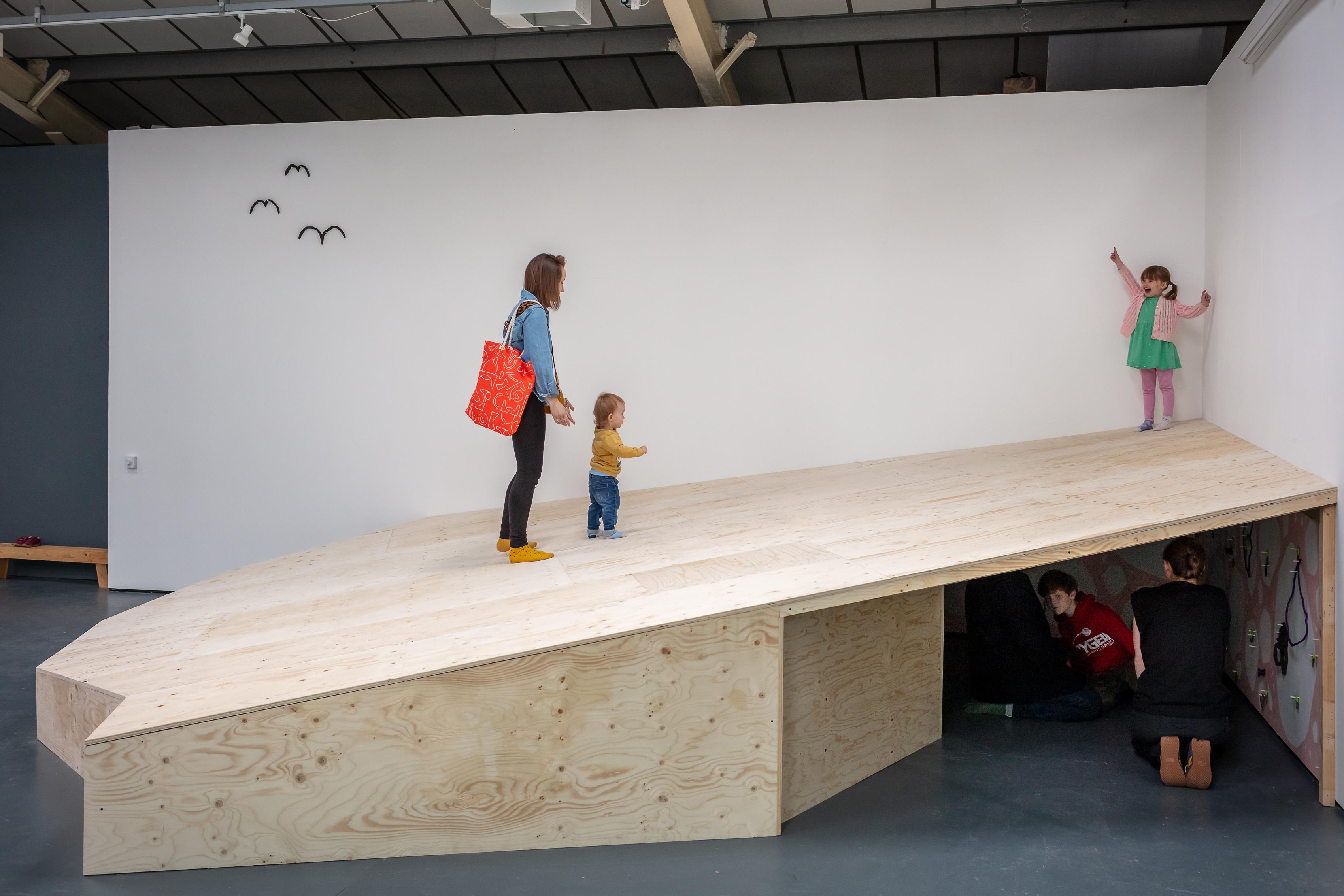 A large wooden ramp fills the corner of the gallery with three figures, an adult and two children on the ramp, and several figures visible in the opening underneath.  The child at the top of the ramp points up, and further along the wall three birds appear to be suspended mid flight as if they were drawn there.