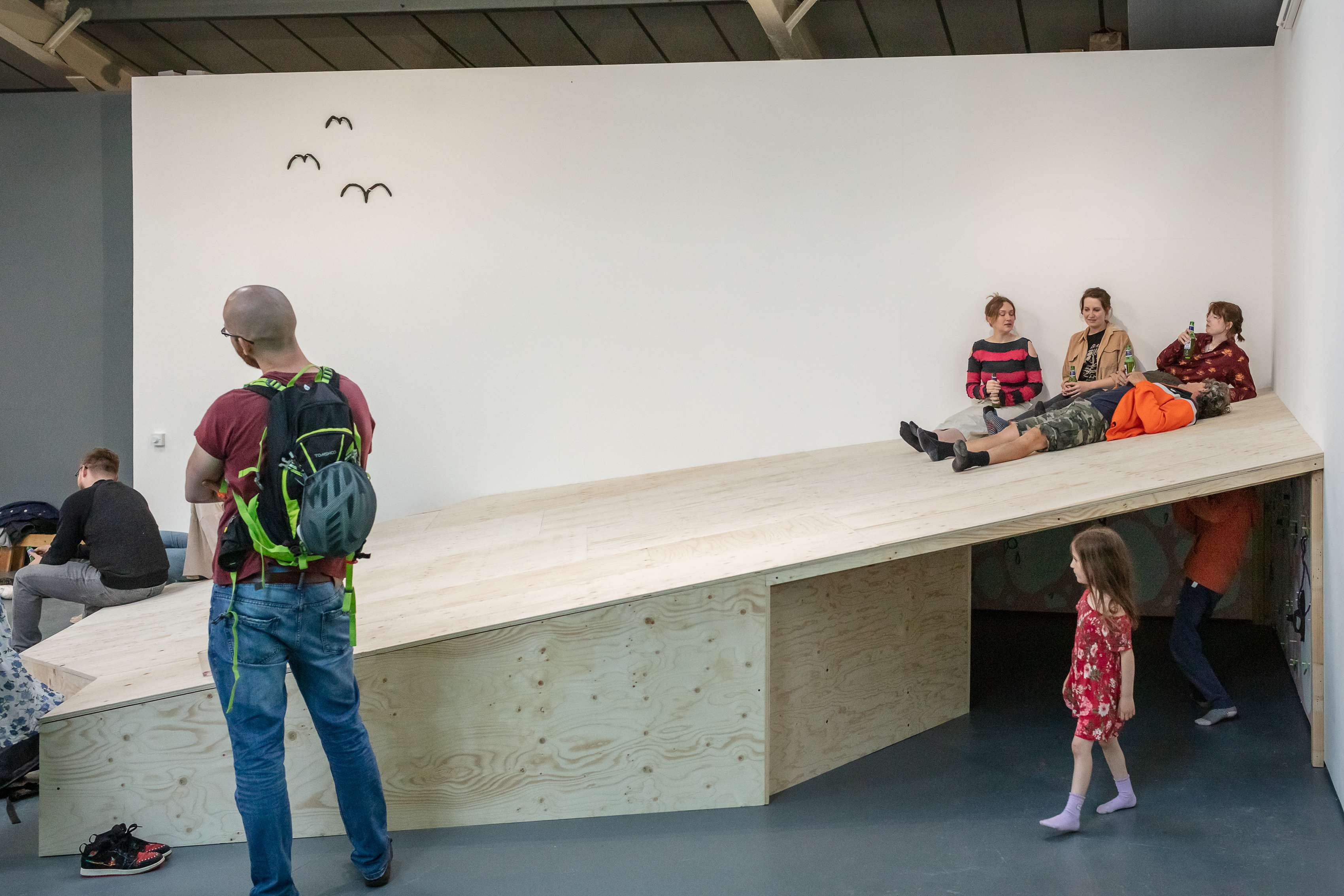 Documentation of hide, a large wooden ramp, being interacted with during the opening of an exhibition. People are seen lying and sitting together at the top, children play underneath, a man stands in front of it and another sits on the edge where it becomes a ledge.