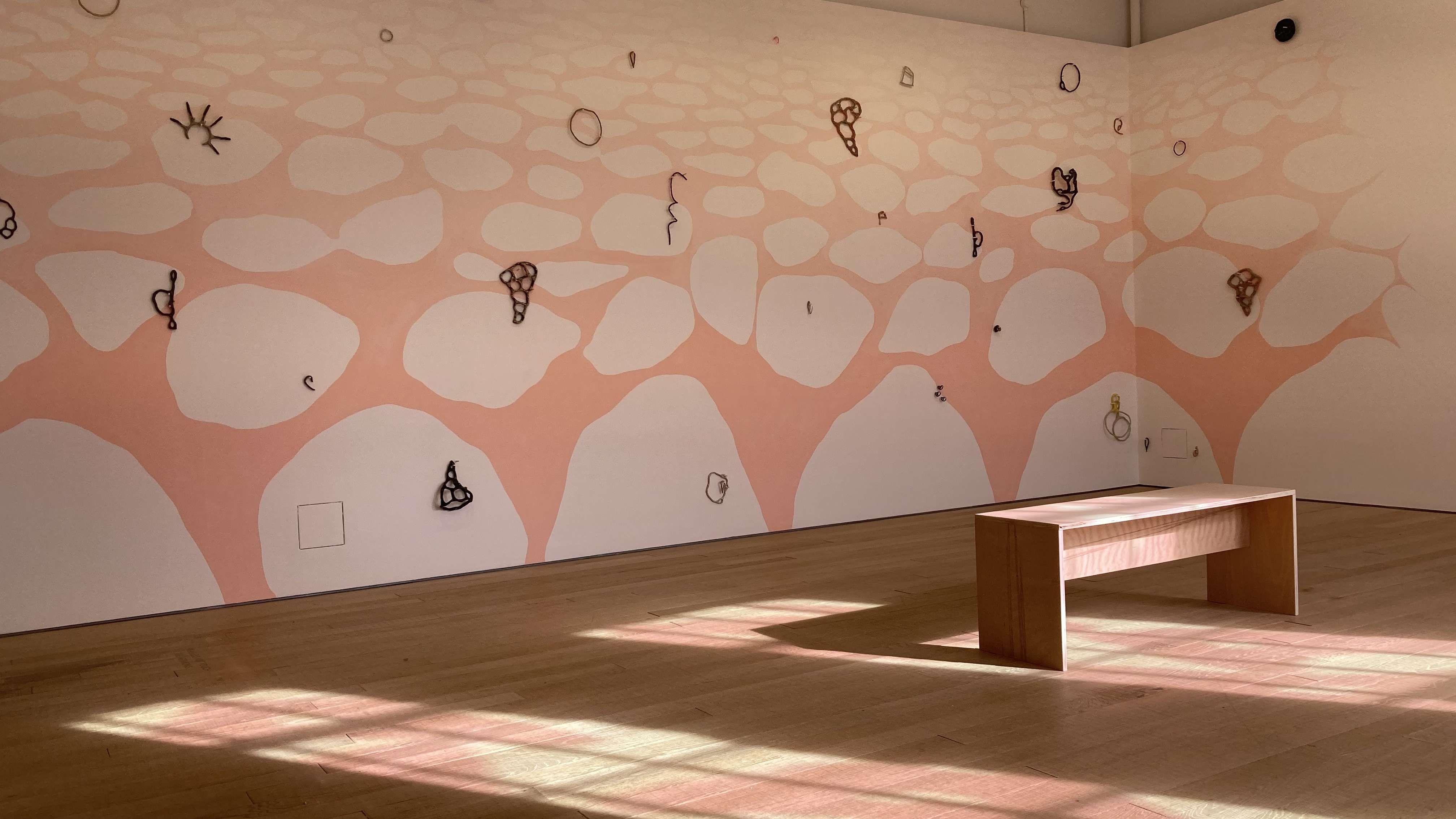 Image shows a wall with white clouds retreating over a pink sky hung with drawn sculptural shapes, with a bench facing it. On the floor blurred pink shapes appear in the bright light of the window shapes.