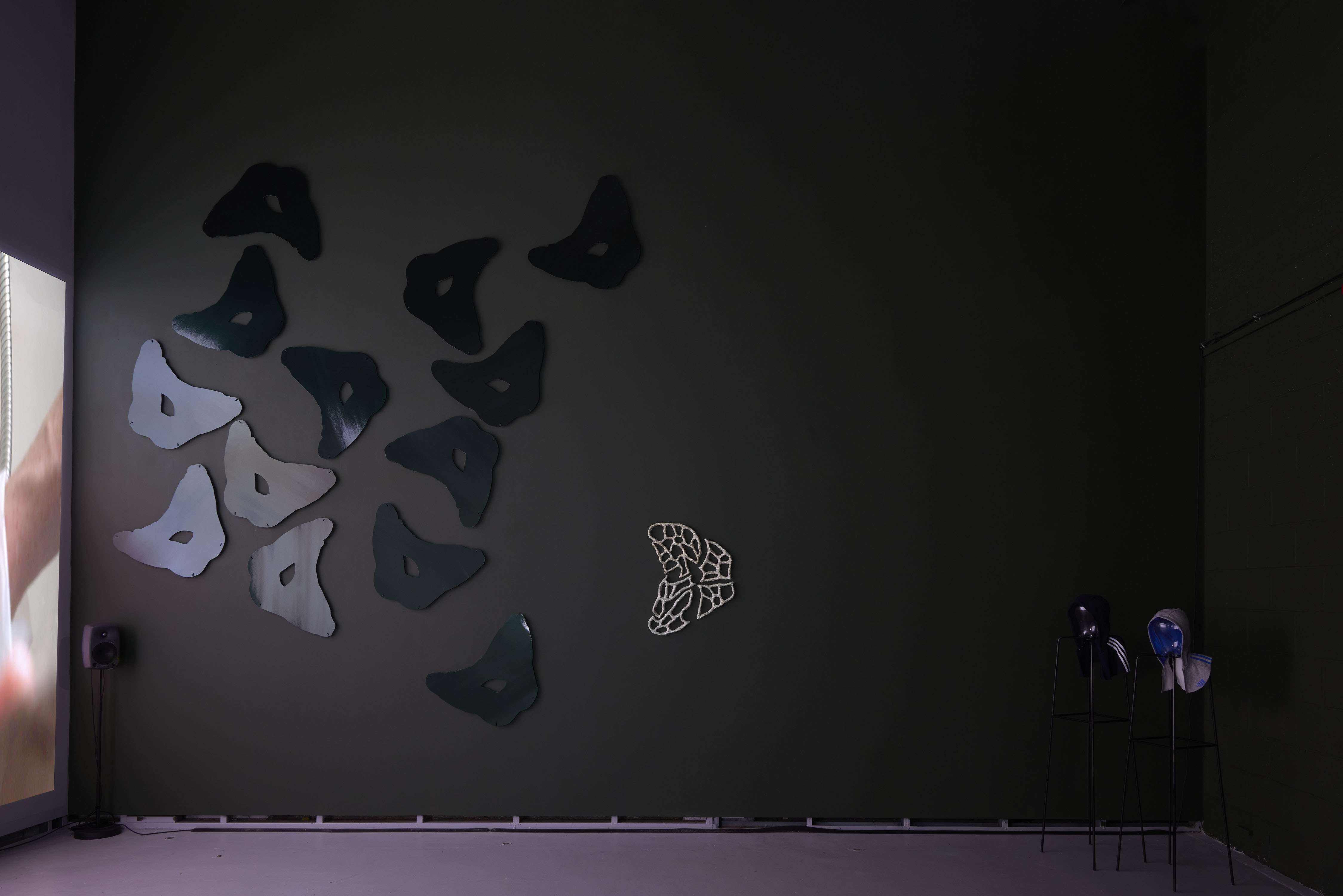 Image shows a dark green wall with several reflective seat shapes reflecting the film that’s playing next to them and mostly out of shot. There is a white webbed sculptural version of the seat in the centre of the wall and in the back corner looking at the film are two figures suggested by glass heads, on black stands, wearing hoods.