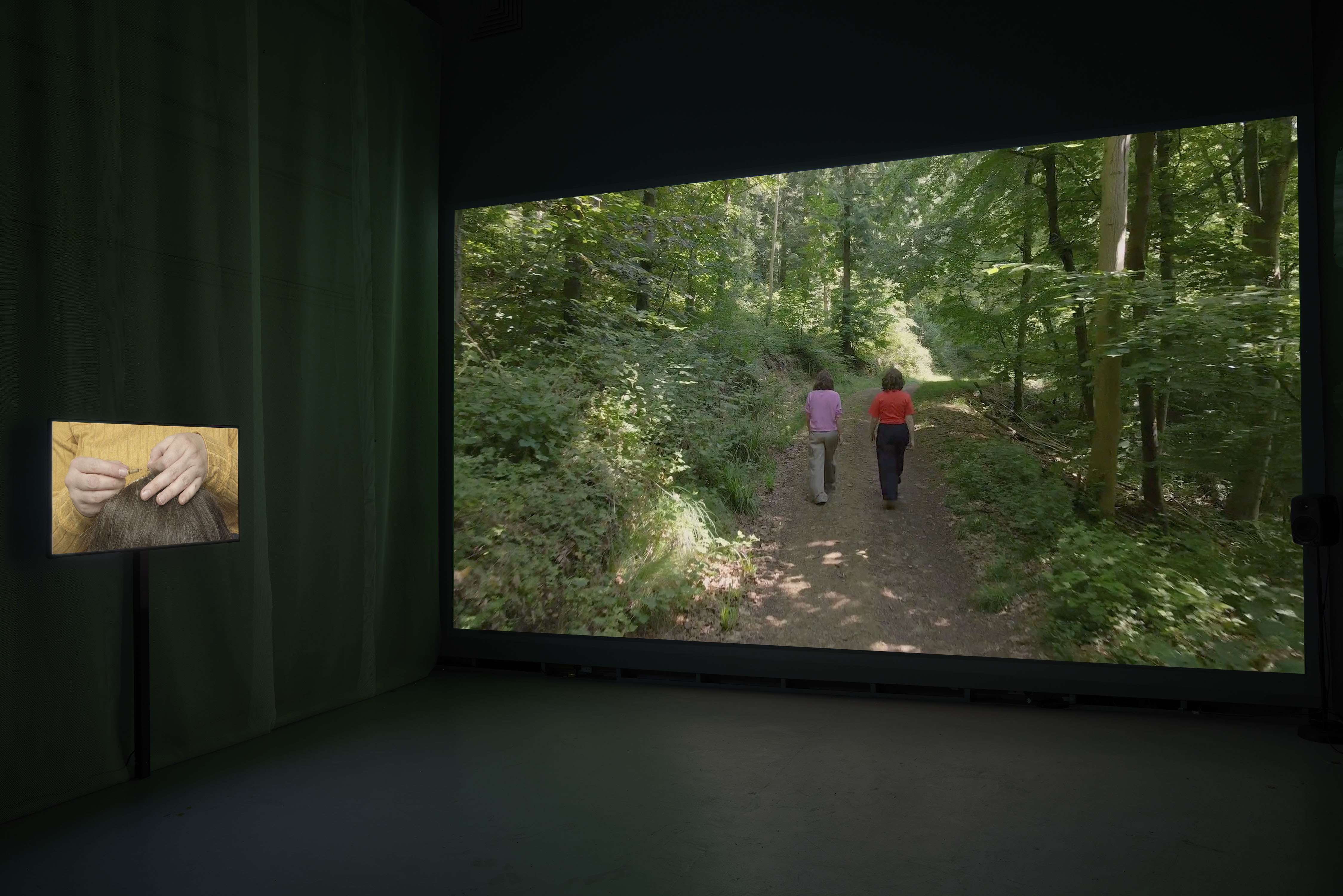 Two films are playing side by side in a dark space. A large film fills the wall showing two figures walking away from the camera in a wood. To its left a monitor is suspended in front of fabric and on screen a pair of hands are knotting hair into a wig.