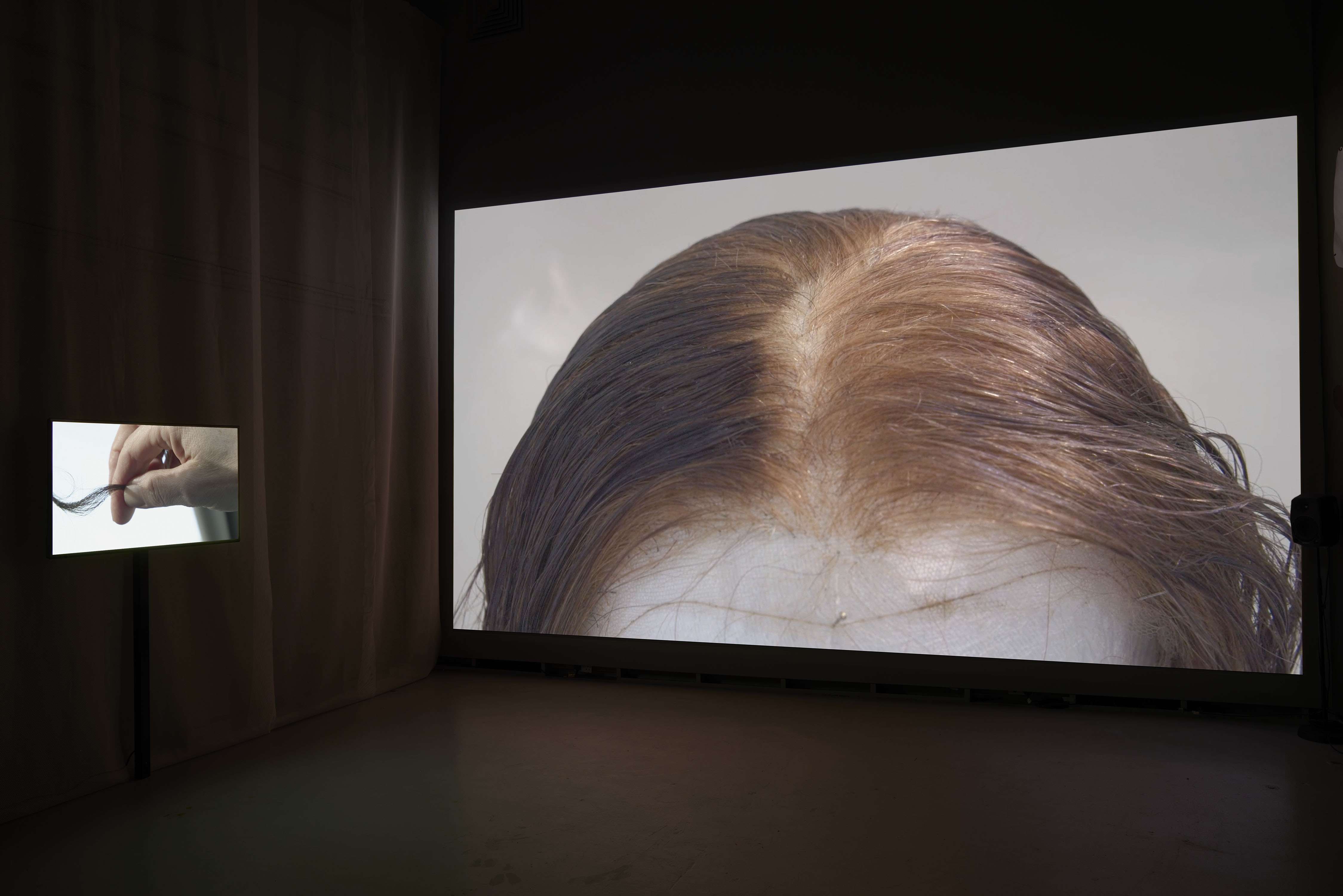 Two films are playing side by side in a dark space. A large film fills the wall showing a wig on a head block with a pin holding it in place. To its left a monitor is suspended in front of fabric and on screen a hand is holding several strands of hair in a twisting motion.