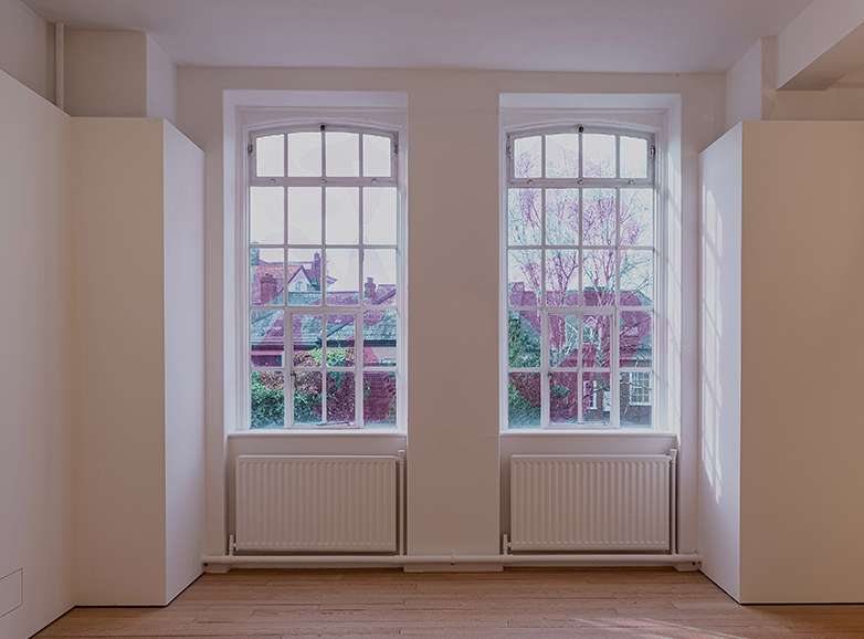 Image shows a gallery space with wooden floors, white walls and two large Victorian windows with radiators below them. In the window panes light pink film, in the shape of someone sitting cross legged on the floor, repeats.
